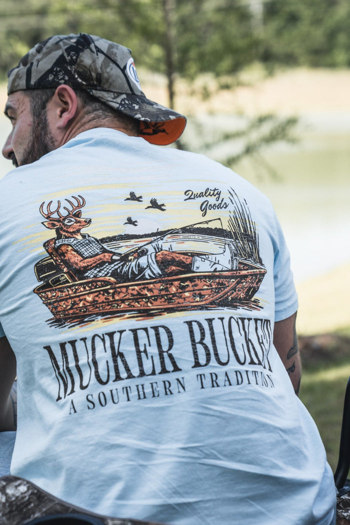 A man relaxes outdoors near a pond and trees in the Buck Dreamin' Premium Tee by Mucker Bucket, enjoying the comfort of this premium cotton fishing t-shirt featuring a deer graphic and “A Southern Tradition” slogan.