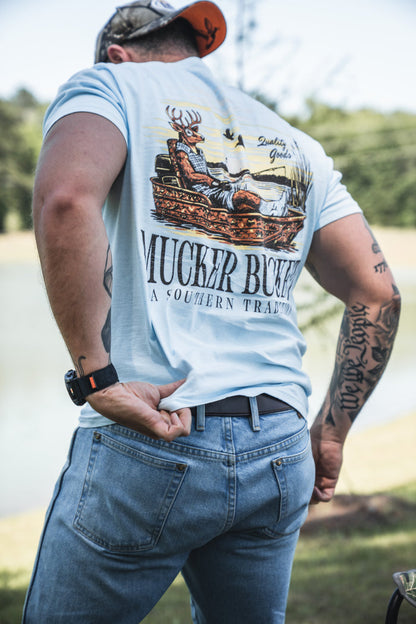 A man in jeans and the Mucker Bucket Buck Dreamin' Premium Tee, featuring a lounging deer and "MUCKIN BUCK, A SOUTHERN TRADITION," adjusts his waistband by a pond. This fishing t-shirt offers premium cotton comfort.