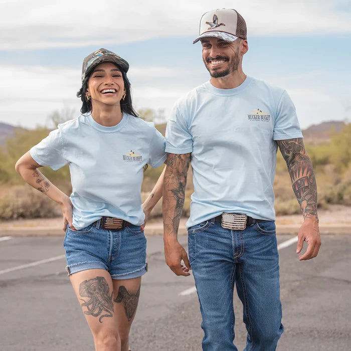 A smiling woman and man wear matching light blue Mucker Bucket Buck Dreamin' Premium Tees, caps, and denim as they walk together outdoors on a sunny day with a desert landscape in the background.