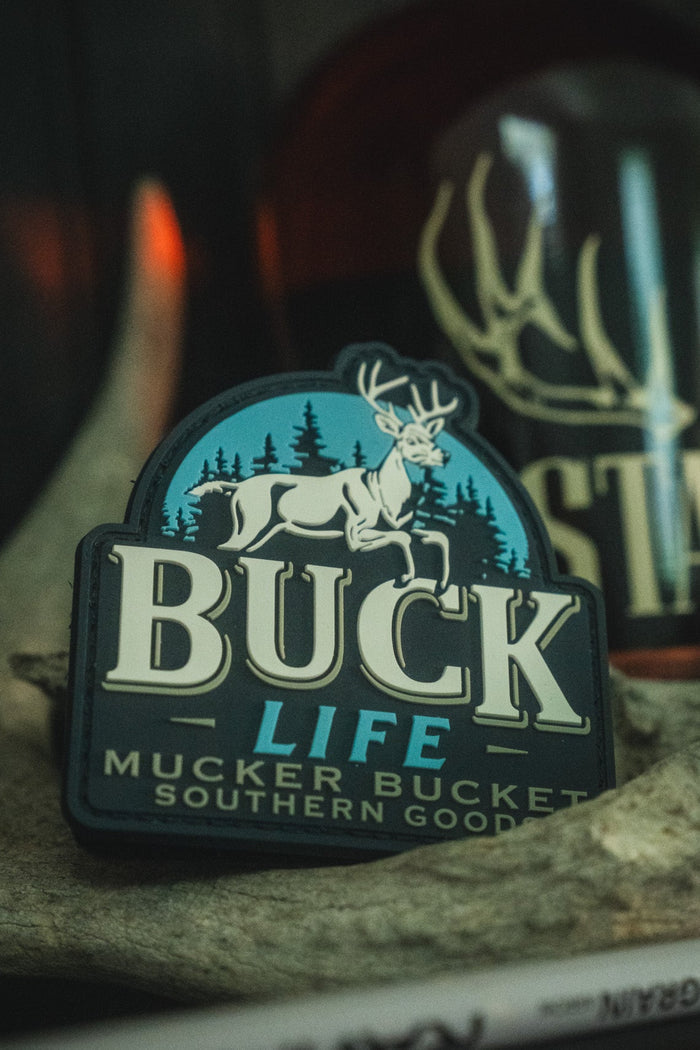 A close-up of a Buck Life patch featuring a white deer on a blue background with the words 'BUCK LIFE' and 'MUCKER BUCKET SOUTHERN GOODS', displayed on a rock with a blurred bottle in the background.