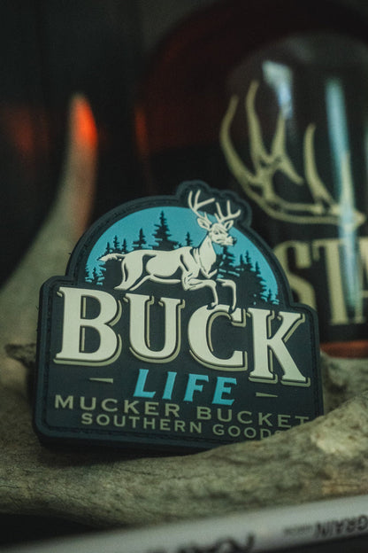 A close-up of a Buck Life patch featuring a white deer on a blue background with the words 'BUCK LIFE' and 'MUCKER BUCKET SOUTHERN GOODS', displayed on a rock with a blurred bottle in the background.