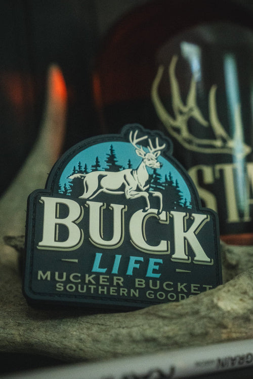 A close-up of a Buck Life patch featuring a white deer on a blue background with the words 'BUCK LIFE' and 'MUCKER BUCKET SOUTHERN GOODS', displayed on a rock with a blurred bottle in the background.
