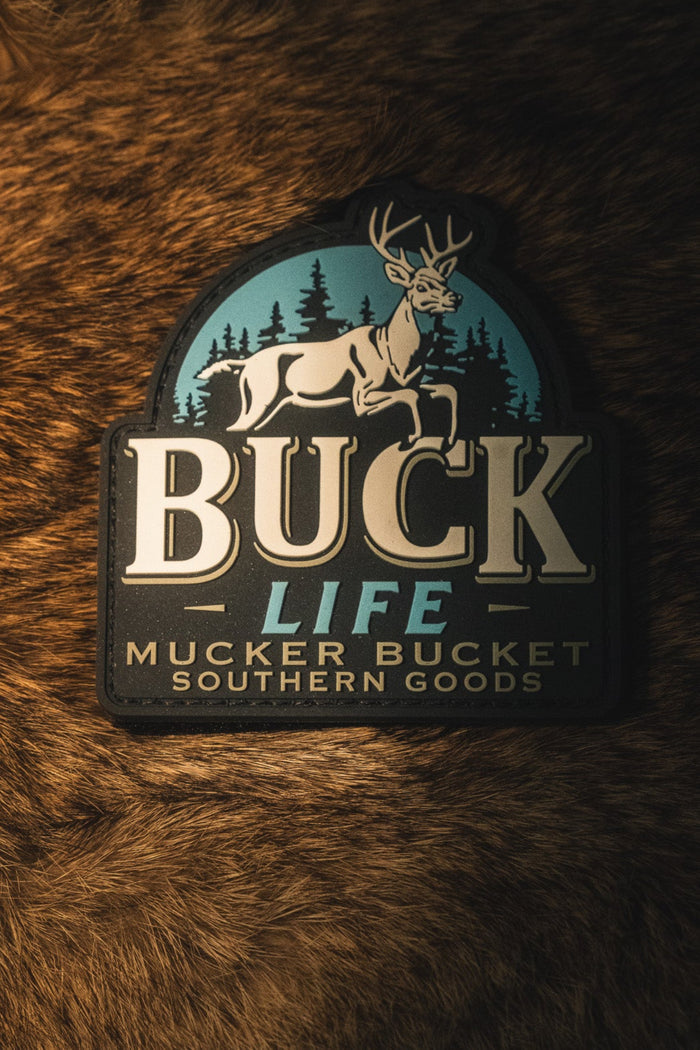 A rectangular patch with rounded top corners featuring a deer and pine trees on a teal background, with the text 'BUCK LIFE MUCKER BUCKET SOUTHERN GOODS' in cream and dark lettering, placed on a brown fur surface.