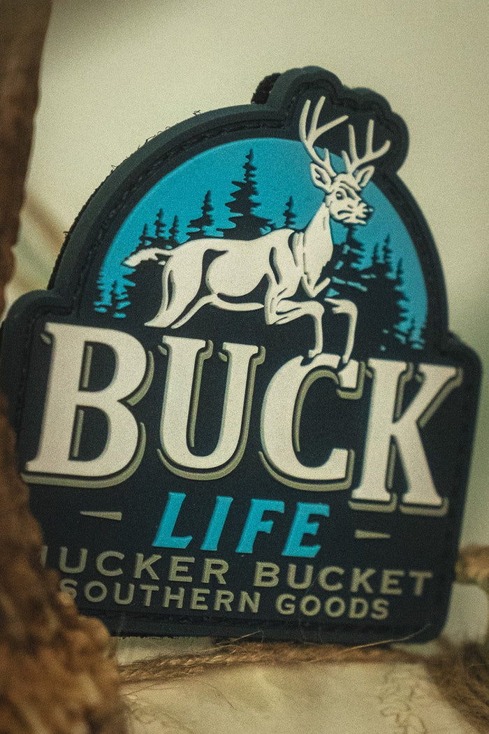 The Buck Life PVC Patch by Mucker Bucket showcases a white deer and trees logo, framed by bold antlers and labeled with "Mucker Bucket Southern Goods," expertly crafted as a durable 4x4 Velcro patch for your favorite gear.