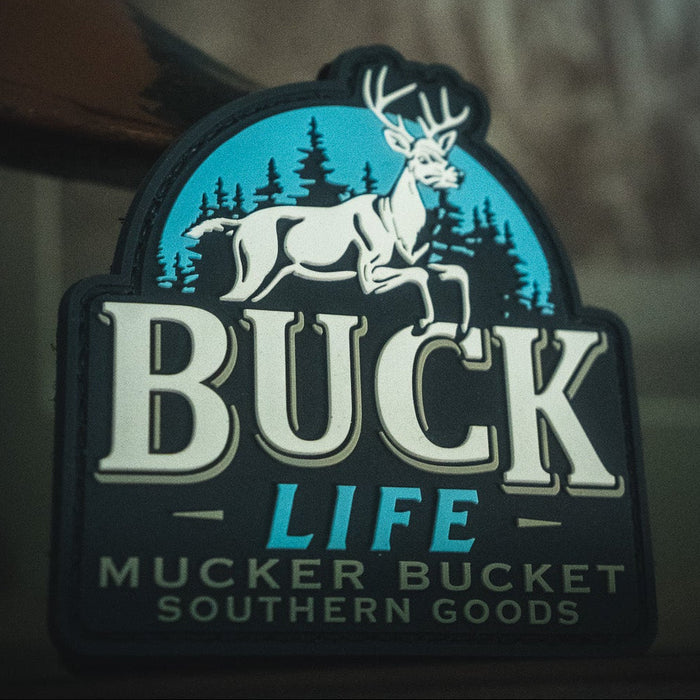 Sticker with a white deer and pine trees reads: Buck Life by Mucker Bucket, perfect for matching with your favorite 4x4 or adding to your Velcro patch collection. Features the bold Buck Life PVC Patch from Mucker Bucket Southern Goods.