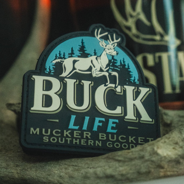 Introducing the Buck Life PVC Patch by Mucker Bucket—a 4x4 inch patch showcasing a white deer, pine trees, and the phrase "Buck Life Mucker Bucket Southern Goods" on a dark background. Crafted from durable 3D PVC and equipped with Velcro backing, it's easy to attach to your favorite gear.