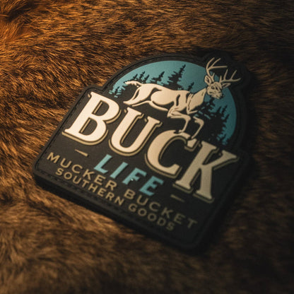 A "Buck Life PVC Patch" from Mucker Bucket, featuring a white deer, trees, and a blue sky in 3D detail, rests on brown fur. The 4x4 patch displays the text “Mucker Bucket Southern Goods” below the scene.