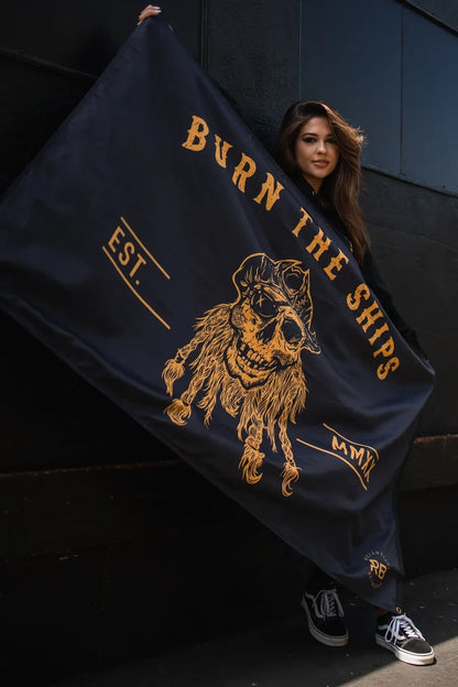 Someone holds the Burn The Ships BLACKOUT Double Sided Flag by Relentless Betrayal, made from premium polyester. It features a bearded pirate skull in gold with Burn the Ships, EST., and MMXIV. They stand casually against a dark wall.