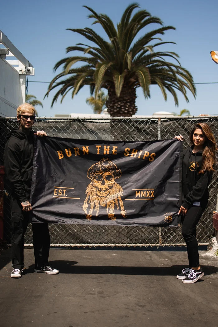 Two people hold a Relentless Betrayal Burn The Ships BLACKOUT Double Sided Flag, made from durable polyester with a stylish pirate skull and EST MMXX text, against a chain-link fence. A palm tree sways under the blue sky as both individuals, in black and sunglasses, stand confidently.