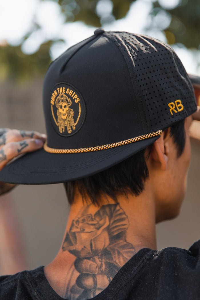 Wearing the Burn The Ships BLACKOUT Snapback from Relentless Betrayal, featuring a sailors emblem and RB on the side, a tattooed individual showcases inked arms with a rose tattoo against a softly blurred tree background.