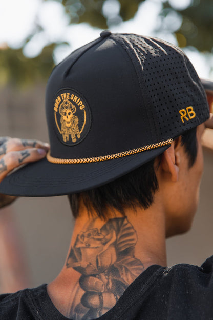 Wearing the Burn The Ships BLACKOUT Snapback from Relentless Betrayal, featuring a sailors emblem and RB on the side, a tattooed individual showcases inked arms with a rose tattoo against a softly blurred tree background.