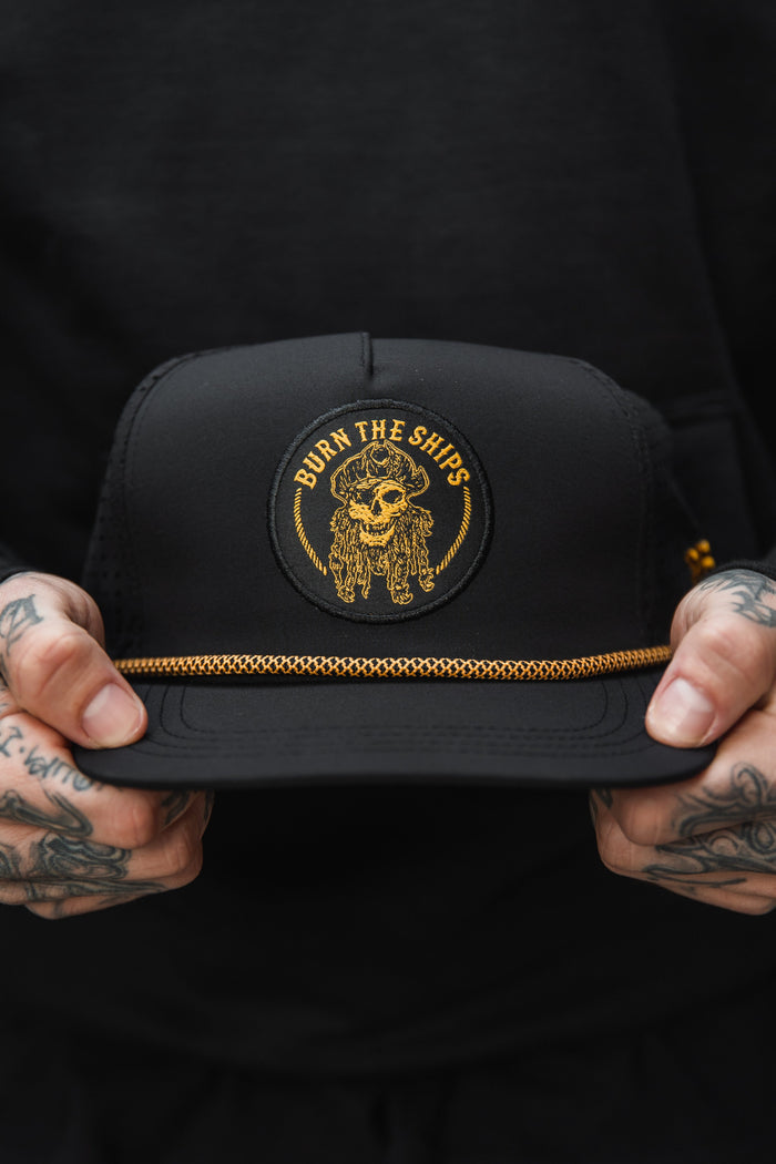 A person with tattooed hands holds the Relentless Betrayals Burn The Ships BLACKOUT Snapback, featuring a circular patch of a skull in a pirate hat with Burn the Ships in gold. A decorative gold chain above the brim completes this unique design.