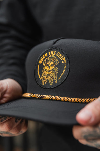 Close-up of a person holding the Relentless Betrayal Burn The Ships BLACKOUT Snapback, featuring a skull pirate patch with Burn the Ships in yellow. This handmade hat includes a decorative yellow rope above the brim, while tattoos adorn the persons fingers.