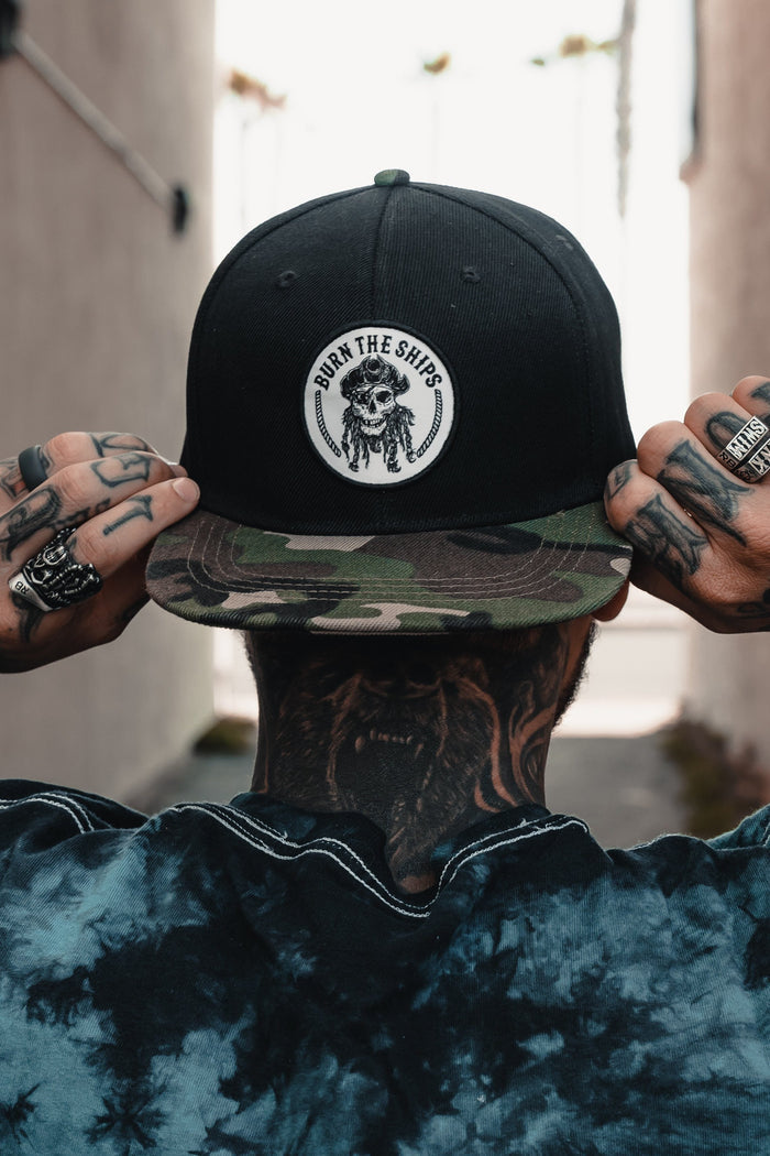With neck and hand tattoos, rings, and a rebellious stance in an alleyway, they wear a Burn The Ships Camo Snapback by Relentless Betrayal. The snapback features a skull and anchor logo that echoes piracys golden era.