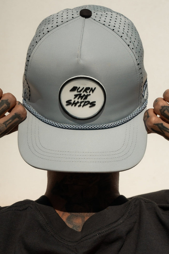 A person dons a Relentless Betrayal Burn The Ships Cement PVC Snapback, embodying the pirate spirit. The light gray cap boasts mesh sides, a textured base, and a water-resistant snapback for durability. Tattooed hands contrast with the plain backdrop.