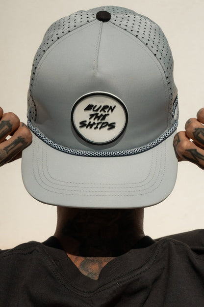 A person dons a Relentless Betrayal Burn The Ships Cement PVC Snapback, embodying the pirate spirit. The light gray cap boasts mesh sides, a textured base, and a water-resistant snapback for durability. Tattooed hands contrast with the plain backdrop.