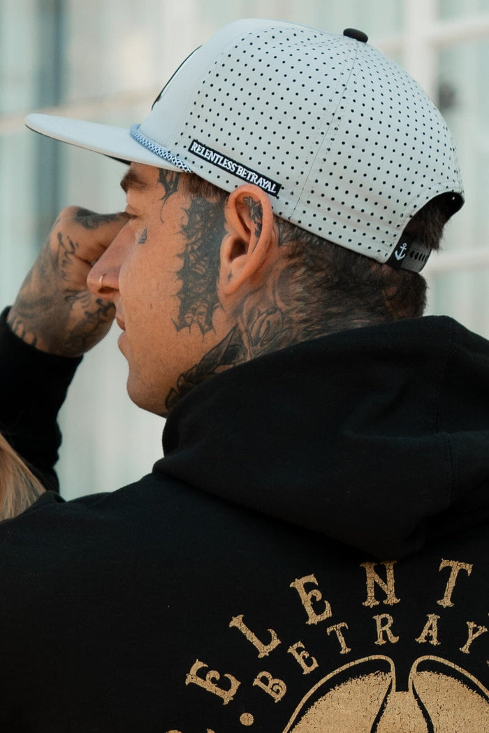 A tattooed individual wears a white Burn The Ships Cement PVC Snapback and a black hoodie with RELENTLESS BETRAYAL on the back, posing outdoors against a sleek glass building.