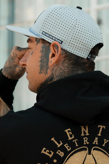 A tattooed individual wears a white Burn The Ships Cement PVC Snapback and a black hoodie with RELENTLESS BETRAYAL on the back, posing outdoors against a sleek glass building.
