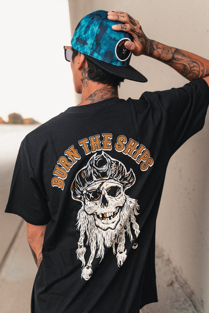 A tattooed individual stands outdoors, leaning against a wall and wearing a backwards blue cap and a Relentless Betrayal black cotton tee featuring BURN THE SHIPS with a pirate skull, hat, and beard.
