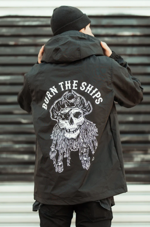 An individual adjusts the hood of their Burn the Ships Pullover Rain Jacket by Relentless Betrayal. The black unisex jacket features a skull in a pirate hat with braids graphic and BURN THE SHIPS above it. Its water-resistant coating stands out against the striped backdrop behind them.