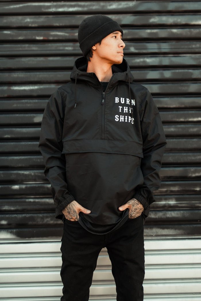 A person in a beanie and hooded jacket stands against a corrugated metal backdrop, looking to the side. The Relentless Betrayal Burn the Ships Pullover Rain Jacket features BURN THE SHIPS on the front, has a water-resistant coating, and pockets for casually tucked hands.