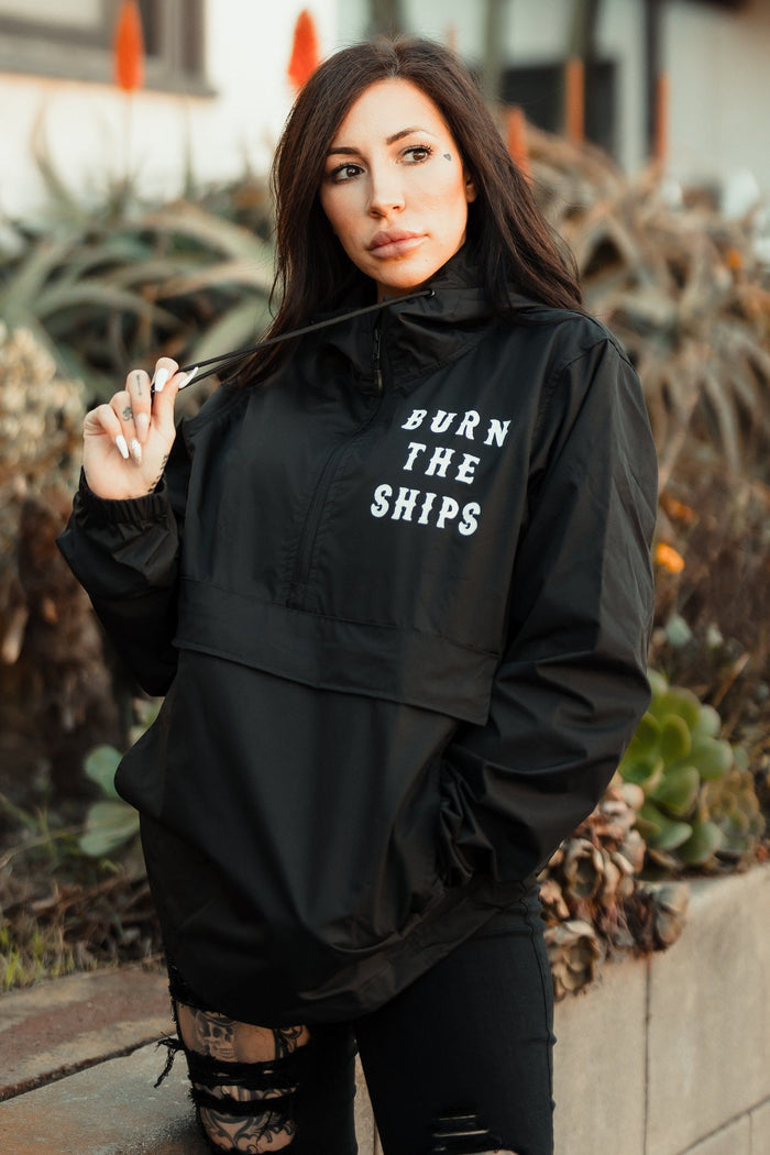 A person with long dark hair stands outdoors wearing the Burn the Ships Pullover Rain Jacket by Relentless Betrayal. This unisex, water-resistant jacket features BURN THE SHIPS on the front. They hold the collar with one hand, surrounded by lush plants.