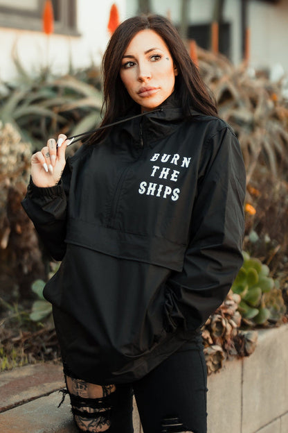 A person with long dark hair stands outdoors wearing the Burn the Ships Pullover Rain Jacket by Relentless Betrayal. This unisex, water-resistant jacket features BURN THE SHIPS on the front. They hold the collar with one hand, surrounded by lush plants.