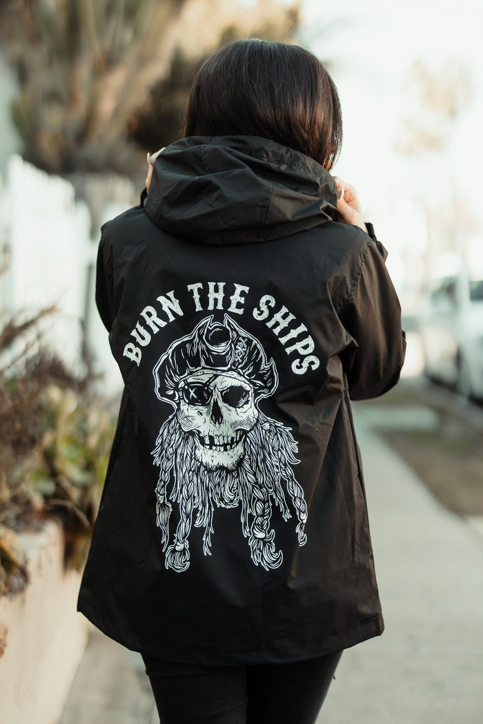 A person wearing the unisex Burn the Ships Pullover Rain Jacket by Relentless Betrayal, featuring a large skull with a pirate hat and BURN THE SHIPS, strides down the sidewalk, framed by blurred plants and cars—perfect for any adventurer.