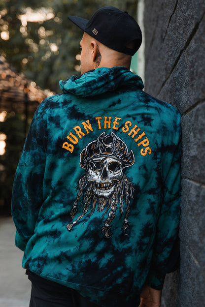 Wearing a Burn The Ships tie-dye hoodie by Relentless Betrayal, featuring a skull design on the back, a man stands against a dark wall with an outdoor blur in the background. Crafted from a cotton-polyester blend for unisex appeal.