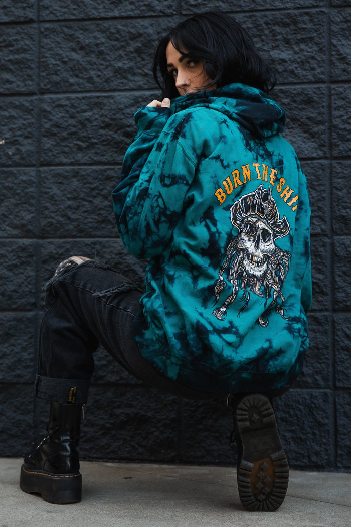 Someone in a Relentless Betrayal Burn The Ships tie dye hoodie, featuring a skull and crossbones design, crouches by a dark brick wall. They wear black ripped jeans and boots, with shoulder-length dark hair.