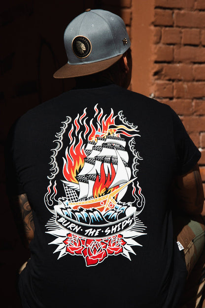 Sporting a Burn The Ships Traditional Tee by Relentless Betrayal, featuring vibrant graphics of a ship with flames and roses and the phrase Burn the Ships, a person wears this unisex fine jersey cotton T-shirt, double-stitched for durability, along with a gray cap in front of a brick wall.