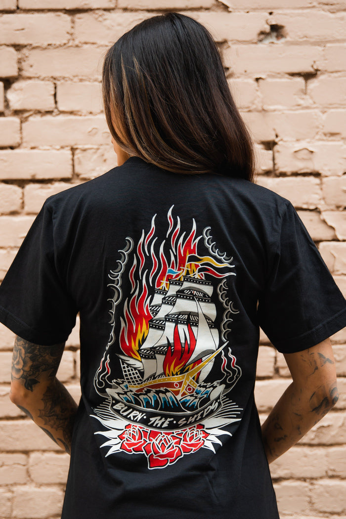 A person with long hair and tattooed arms faces away, wearing the Relentless Betrayal Burn The Ships Traditional Tee, featuring a flaming ship with red roses. Made from fine jersey cotton with a double-stitched design, it stands out against a light brick wall.