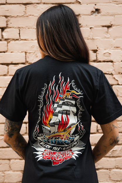 A person with long hair and tattooed arms faces away, wearing the Relentless Betrayal Burn The Ships Traditional Tee, featuring a flaming ship with red roses. Made from fine jersey cotton with a double-stitched design, it stands out against a light brick wall.