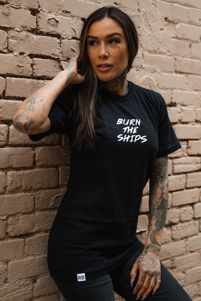 A tattooed person stands against a brick wall wearing the Burn The Ships Traditional Tee by Relentless Betrayal. The unisex black T-shirt with double-stitched fine jersey cotton features BURN THE SHIPS. Their long hair flows as one hand casually touches their head.