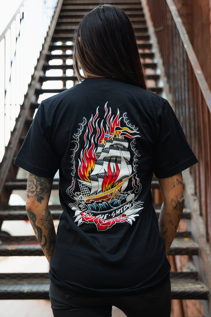 Standing on a staircase with long hair and tattoos, the individual sports a Relentless Betrayal Burn The Ships Traditional Tee, a black unisex fine jersey cotton shirt featuring a vibrant ship-in-flames graphic and double-stitched design.