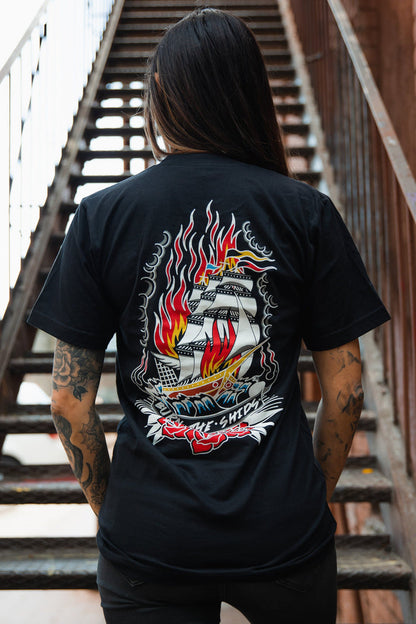 Standing on a staircase with long hair and tattoos, the individual sports a Relentless Betrayal Burn The Ships Traditional Tee, a black unisex fine jersey cotton shirt featuring a vibrant ship-in-flames graphic and double-stitched design.