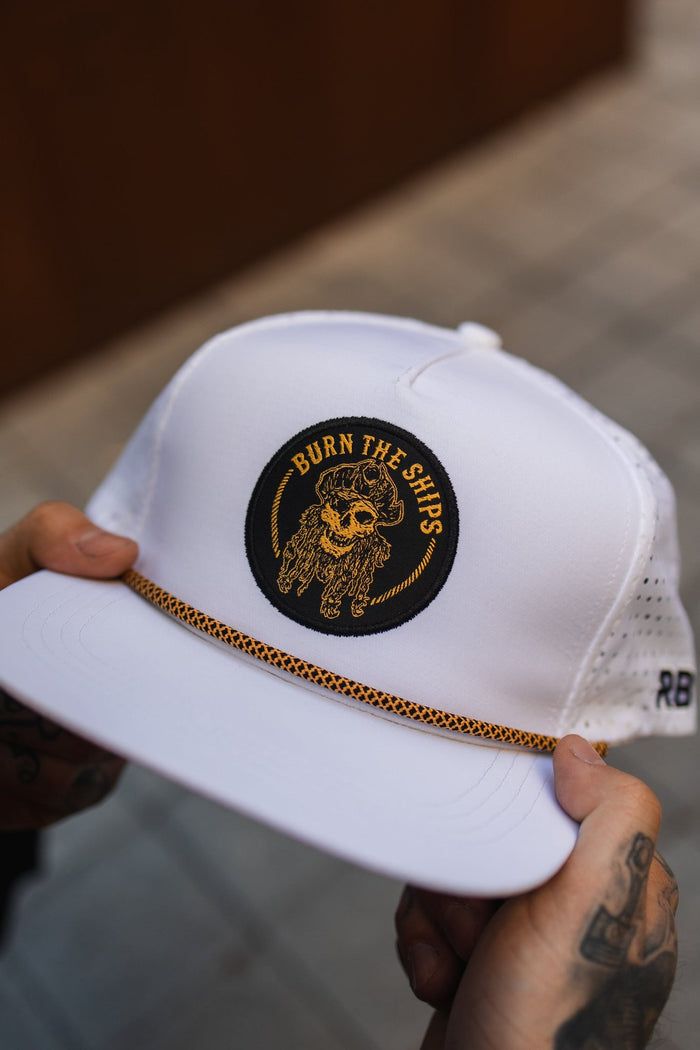 Two hands hold a water-resistant Burn The Ships WHITEOUT Snapback by Relentless Betrayal, featuring a circular black patch with an embroidered pirate skull and BURN THE SHIPS in gold. The hat has a perforated back and a decorative gold band above the brim.