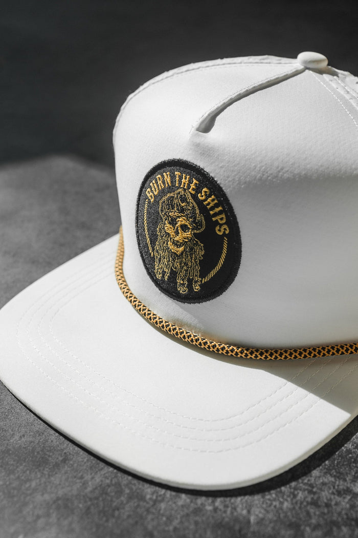 The Burn The Ships WHITEOUT Snapback by Relentless Betrayal features a white cap with a black and gold embroidered pirate patch of a bearded skull with BURN THE SHIPS. It includes a gold braided cord on this water-resistant hat, displayed on a textured gray surface.