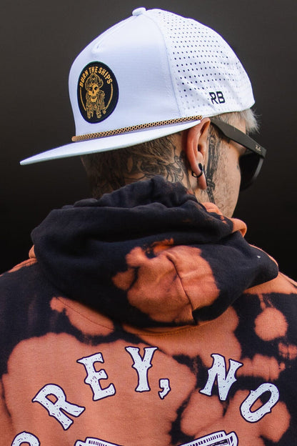 A tattooed individual wears a Relentless Betrayal Burn The Ships WHITEOUT snapback and sunglasses, paired with a black and orange tie-dye hoodie featuring OBEY, NO, facing away from the camera.