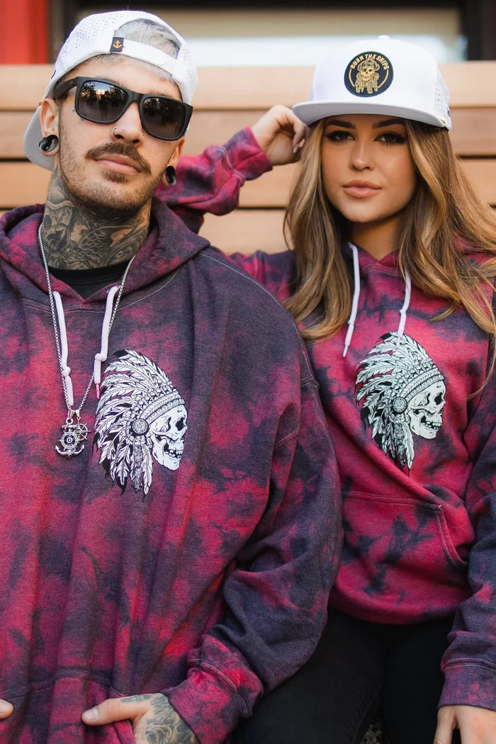 Two people in matching red and black tie-dye hoodies with a skull headdress design sit on wooden steps. The man wears sunglasses and a snapback, while the woman dons the Relentless Betrayal Burn The Ships WHITEOUT Snapback with an embroidered pirate patch.