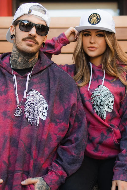 Two people in matching red and black tie-dye hoodies with a skull headdress design sit on wooden steps. The man wears sunglasses and a snapback, while the woman dons the Relentless Betrayal Burn The Ships WHITEOUT Snapback with an embroidered pirate patch.