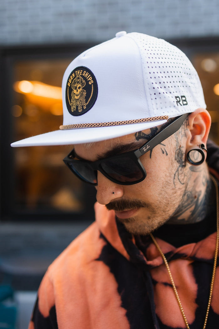Wearing a Burn The Ships WHITEOUT Snapback by Relentless Betrayal with an embroidered pirate patch, black sunglasses, and a tie-dye hoodie, a tattooed person with large earrings looks down in an outdoor setting against a blurred background.
