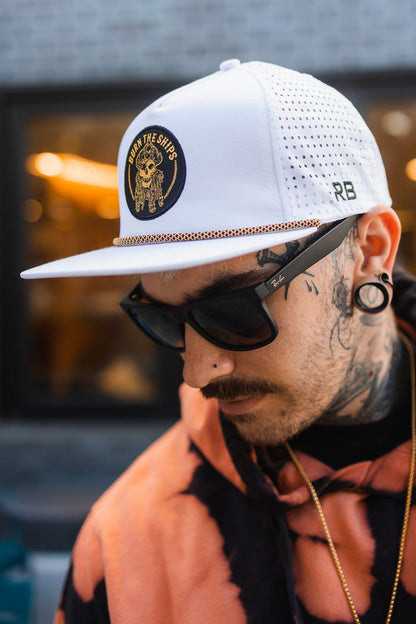 Wearing a Burn The Ships WHITEOUT Snapback by Relentless Betrayal with an embroidered pirate patch, black sunglasses, and a tie-dye hoodie, a tattooed person with large earrings looks down in an outdoor setting against a blurred background.