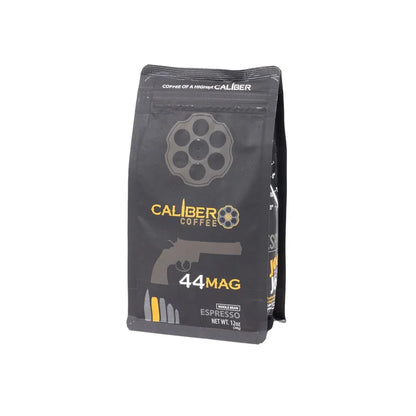 A black package of Caliber Coffee Company features gun-themed graphics, including a revolver chamber and handgun silhouette. The label on Caliber Espresso Roast .44 Mag - Whole Bean displays a net weight of 12 oz, highlighting artisan roasted, 100% Arabica beans for specialty grade coffee lovers.