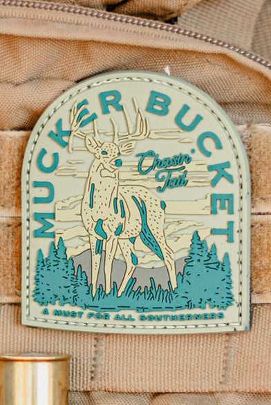 The tan backpack features the Mucker Bucket "Chasin' Tail 2.O PVC Patch," displaying a deer in a forest with mountain backdrop. This blue and cream patch is perfect for Southerners seeking standout outdoor-inspired style.