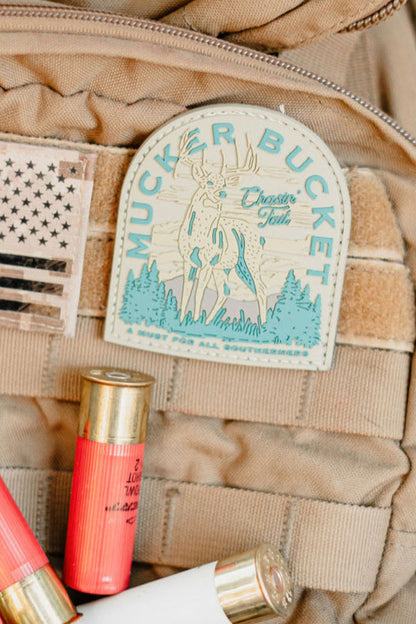 A close-up of a tan tactical vest showcases the Chasin' Tail 2.O PVC Patch by Mucker Bucket, featuring a deer illustration, alongside a distressed American flag patch and several red and white shotgun shells—ideal for outdoor enthusiasts.