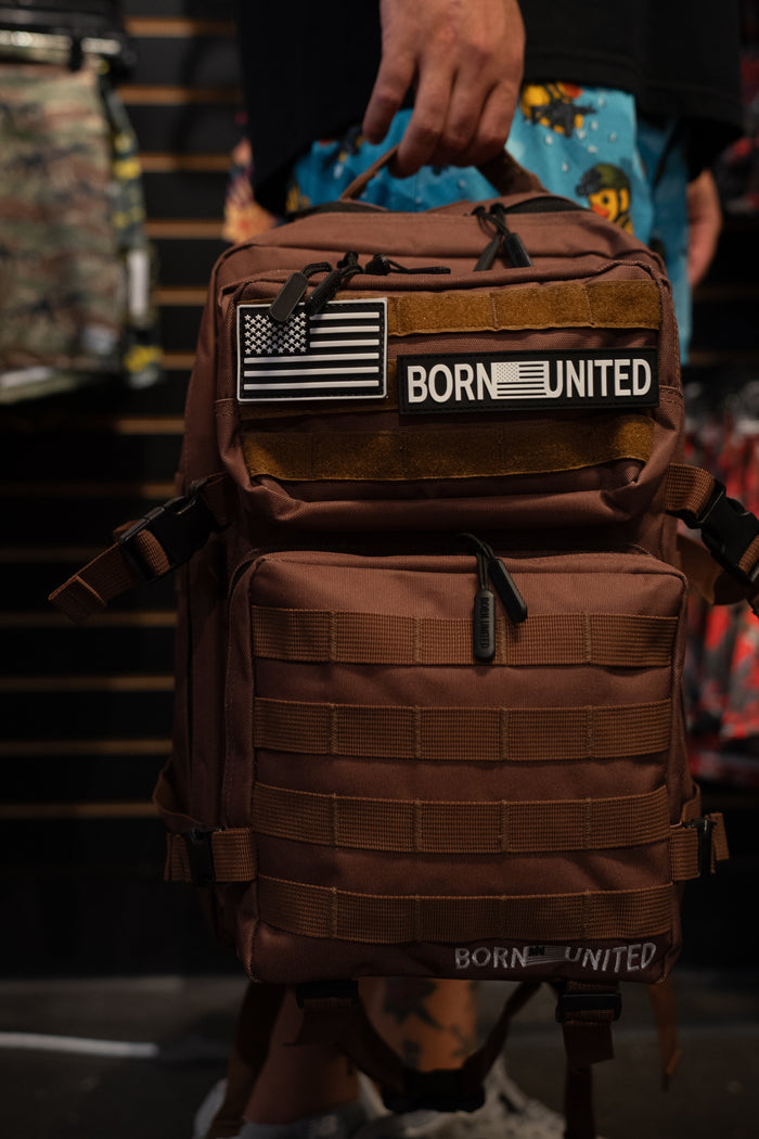 A person holds the Chocolate Tac-Pac - 25L by Born United Private Label, featuring black and white American flag and BORN UNITED patches, while standing in a store. Designed for organized storage, this tactical backpack seamlessly blends functionality with style.