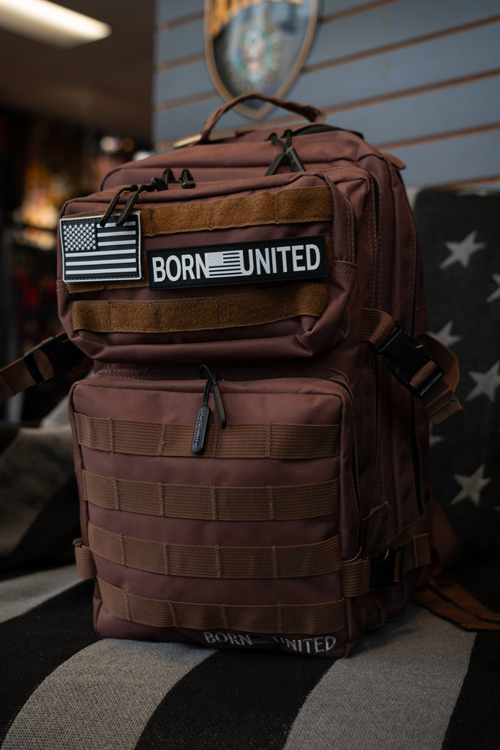The Chocolate Tac-Pac - 25L by Born United Private Label, adorned with American flag and BORN UNITED patches, features organized storage and is displayed indoors on a star-patterned chair.