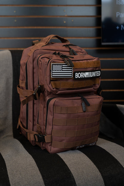 The Born United Private Label Chocolate Tac-Pac - 25L, featuring organized storage, multiple pockets, a U.S. flag patch, and a BORN UNITED patch, sits on a striped chair indoors.