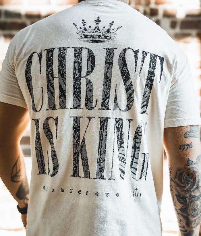 Someone wears the Christ is King Premium Tee - Ivory by 13th, with a crown above bold black "CHRIST IS KING" text and smaller "THIRTEEN 13TH" below, set against a blurred brick background.